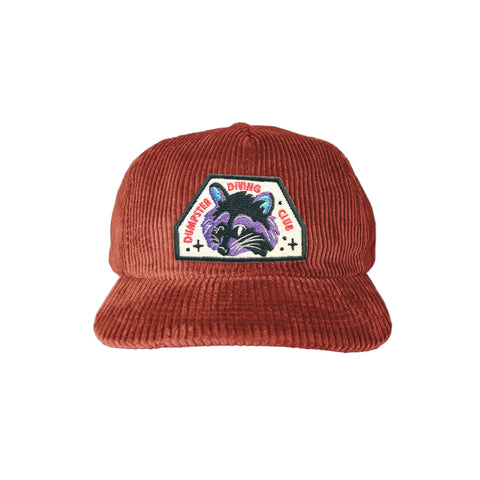 Brick red corduroy hat. It has a raccoon patch that reads "dumpster diving club"