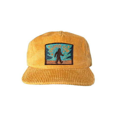 Mustard-colored 5 panel corduroy hat with a bigfoot patch that reads "cryptid club"