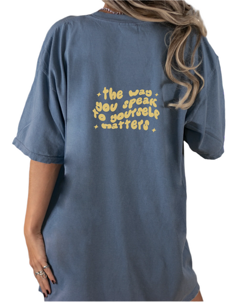 The back view of a woman wearing a blue shirt with yellow text that reads "the way you speak to yourself matters"