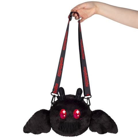 A hand holding a black crossbody bag with a plush design resembling a baby mothman with red eyes and magnetic wings.