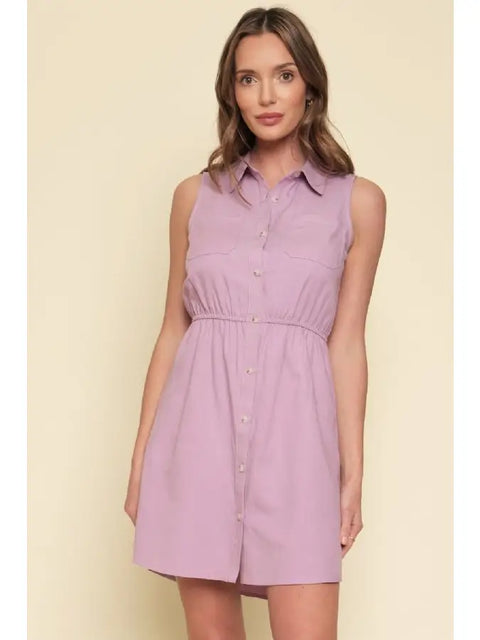 Woman wearing a sleeveless purple dress against a beige background. It is button down and has a cinch waist.