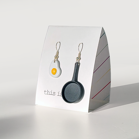 Image shows a pair of earrings hanging from silver wires. One is a frying pan, and one is a fried egg.