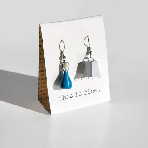 Image shows a lamp-themed pair of earrings hanging from silver wires. The base is blue and the shade is white.