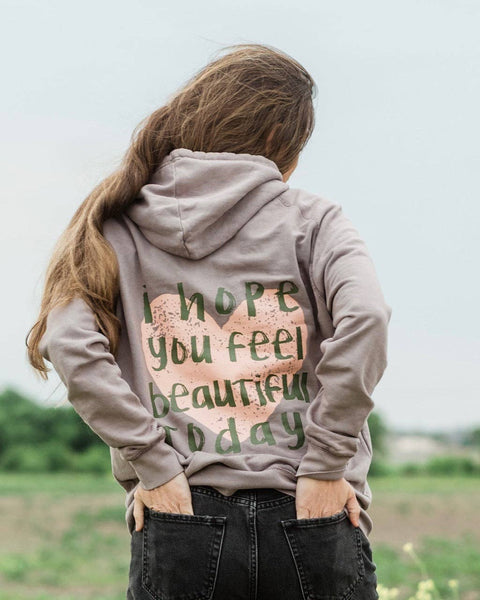A woman standing in a grassy field wearing a grey sweatshirt with the text "I hope you feel beautiful today" over a heart