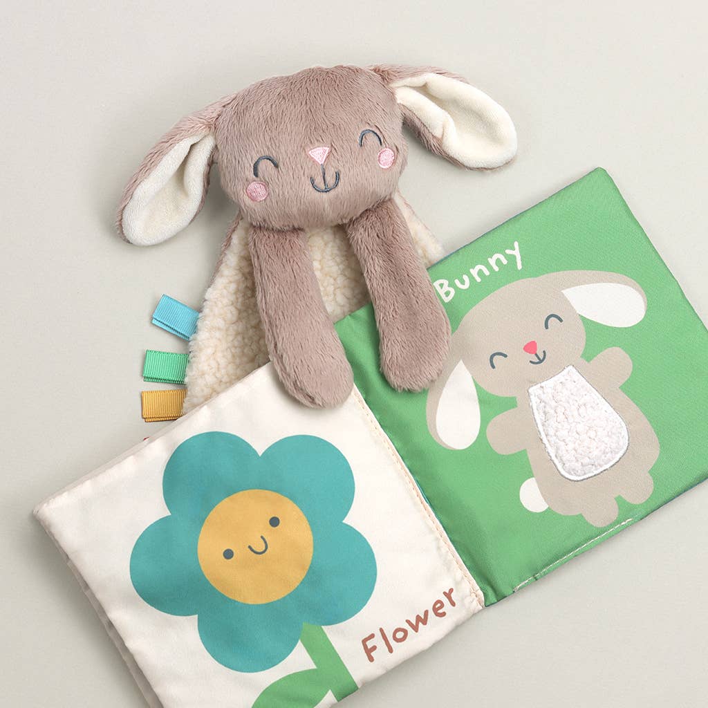 Image shows a bunny lovey holding open a fabric book with a flower on one page and a bunny on the other