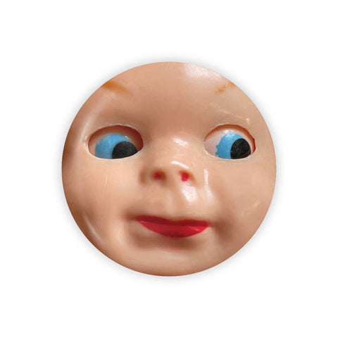 Image shows a round magnet with a plastic doll face. It has blue eyes that are looking to the side.