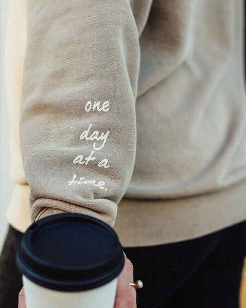 A woman holding a to go coffee cup wearing a sand colored sweatshirt that reads "one day at a time" near the wrist