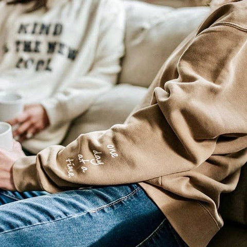 Two women sitting together. The woman in the foreground wears a sand colored sweatshirt with the text "one day at a time" on the sleeve.