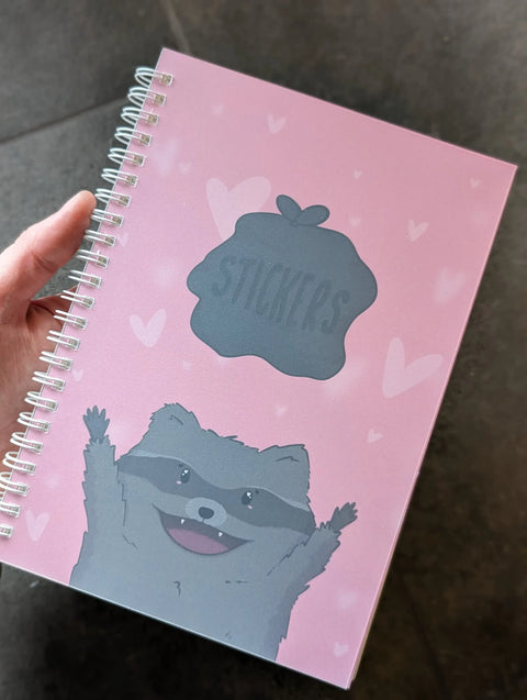 Image shows a pink spiral bound sticker book. It features a racoon throwing a bag of trash that reads "stickers"