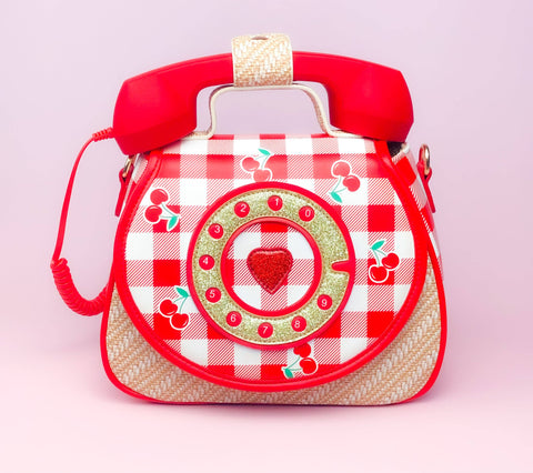 Image shows a handbag with a red phone on top. It has red checkerboard with cherries and a gold rotary dial.