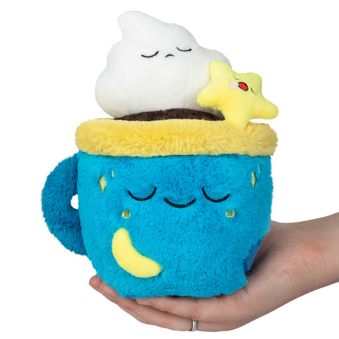 A plush toy resembling a blue coffee mug with whipped cream, yellow stars, and a brown liquid resin handle, held in a hand. The cup and whipped cream have sleepy faces.