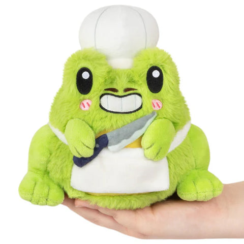 A green frog plush toy wearing a chef's outfit, including a hat and apron, held in a person's hand.