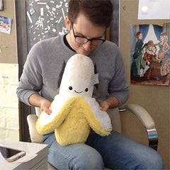 A man holds the plush banana in his lap. It is about the size of his torso.