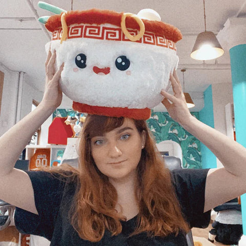 A woman balances the plush ramen bowl on her head. It is significantly larger than her head. 