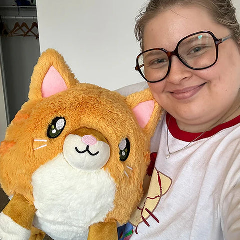 A woman holds the stuffed orange cat. It is slightly larger than her chest.