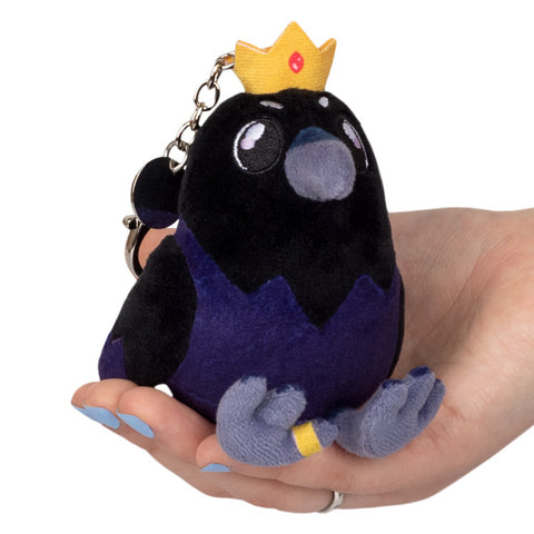 A plush toy resembling a crow wearing a crown, held in a person's hand. It is attached to a silver clip keychain.