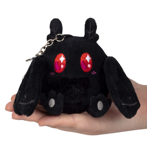 A small black plush toy resembling a mothman with red and pink eyes, being held in a person's hand. It is attached to a silver clip keychain.