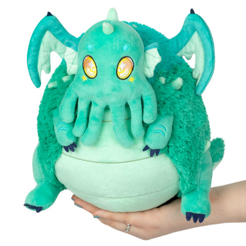 A plush toy resembling a baby Cthulhu with a predominantly green color, held in someone's hand.