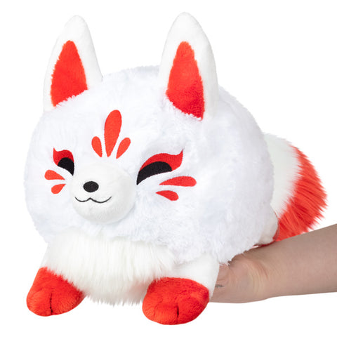 A plush toy resembling a Kitsune with a white body, red markings, black eyes with red accents, and red feet and tail tips. It is being held in a person's hand.