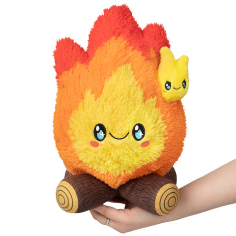 A plush toy resembling a mini campfire with a smiling face, in shades of yellow and orange, held in someone's hand.