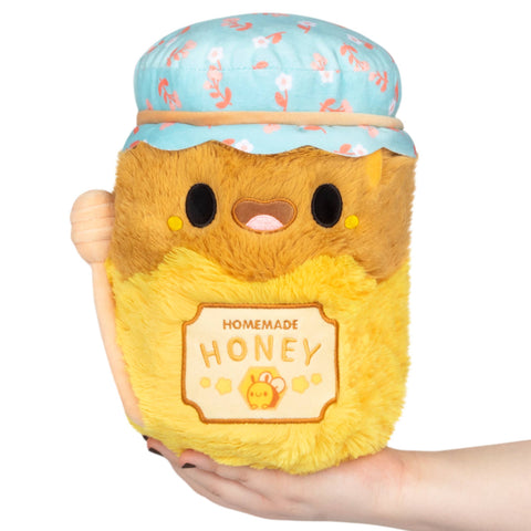 Plush toy shaped like a honey jar with a blue lid held by a hand on a white background