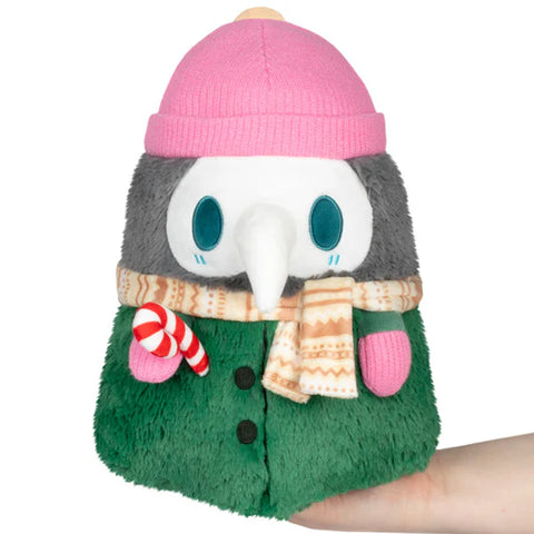 Image shows a green and grey stuffed plague doctor wearing a pink beanie and gloves and tan scarf.
