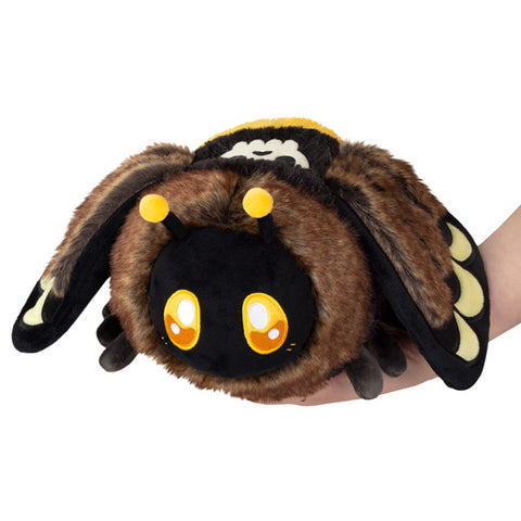 A plush toy resembling a Deaths-Head Hawkmoth moth with brown wings, black body, and yellow eyes, being held in a person's hand.