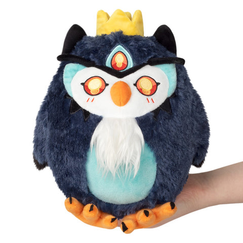 A plush toy depicting a mini demon owl with a blue body, black wings, a yellow crown, and orange feet. It is held in someone's hand.