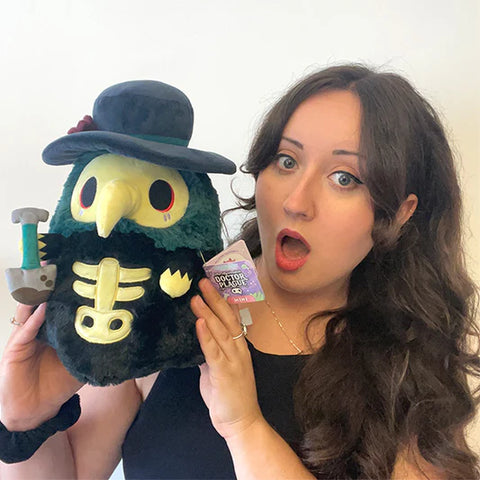 Woman holding the plush graveyard-themed plague doctor. It is slightly larger than her head.