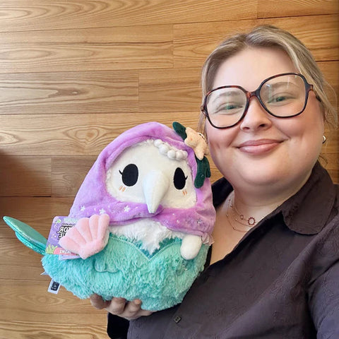 A woman holds the stuffed mermaid plague doctor. It is slightly larger than her head.