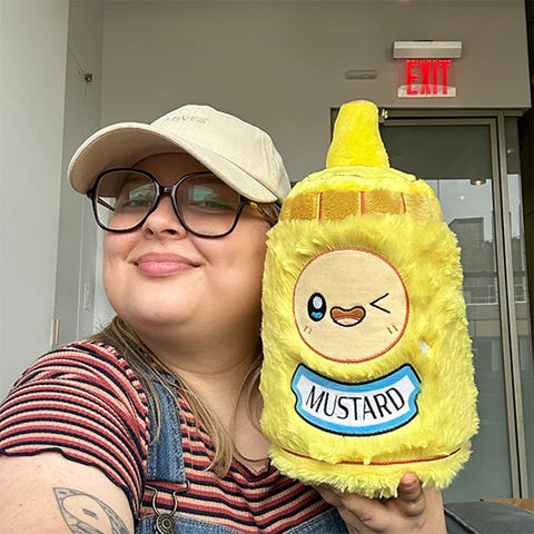 Image shows a woman holding the stuffed mustard bottle up to her face. It is slightly larger than her head.