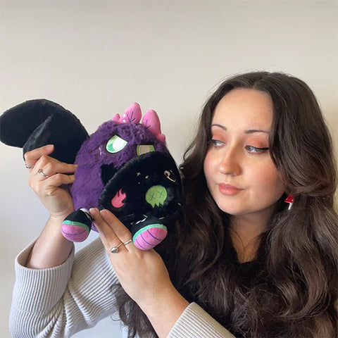 A woman holds the plush neon punk baby mothman near her face. It is slightly larger than her head.