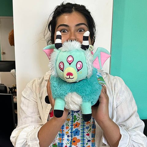A woman holds the plush pastel demon in front of her face. It is slightly larger than her head.