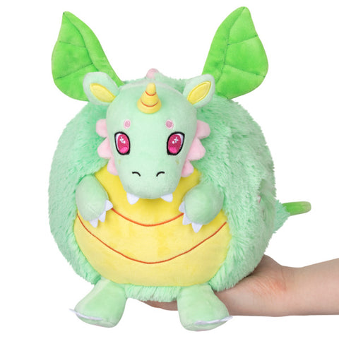 A plush toy resembling a green dragon with a yellow belly and horn, and pink accents, such as cheeks and inner mouth, held in a person's hand.