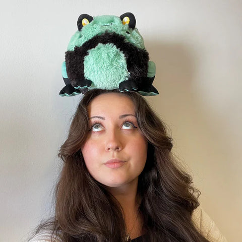 A woman balances the plush frog on her head. It is slightly larger than her head.