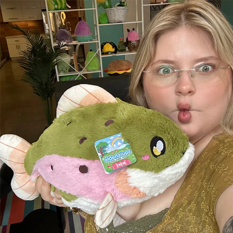 A woman makes a fish face while holding the plush rainbow trout. It is slightly larger than her head.