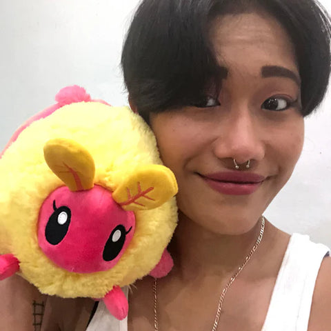 A woman holds the plush rosy maple moth up to her face. It is slightly larger than her head.