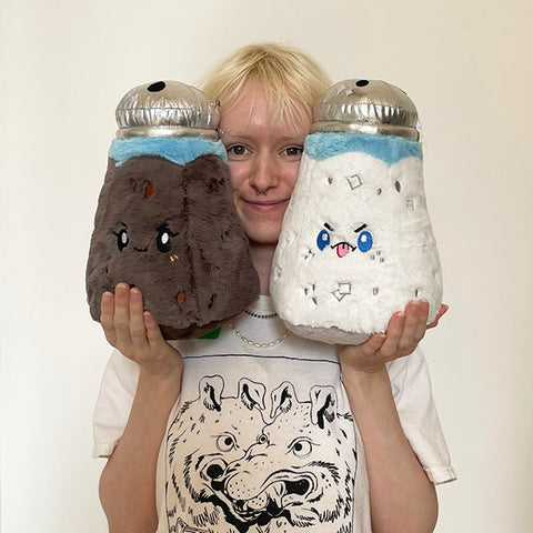 A woman holds the plush saltshaker and a plush peppershaker up to her face. They are slightly larger than her head.