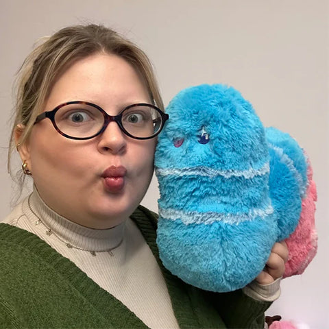 A woman holds the plush sour gummy worm near her face. It is slightly larger than her head.