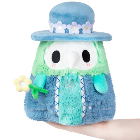 Image shows the stuffed spring plague doctor. He is pastel blue and green, has a daisy chain around his hat, and is holding a flower.