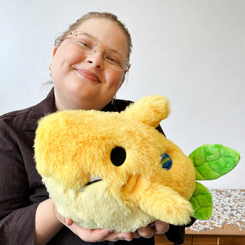 A woman holds the plush lemon shark near her body. It is about the size of her chest.