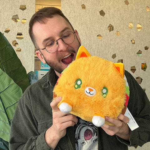 A man pretends to take a bite out of the plush taco cat. It is slightly larger than his head.