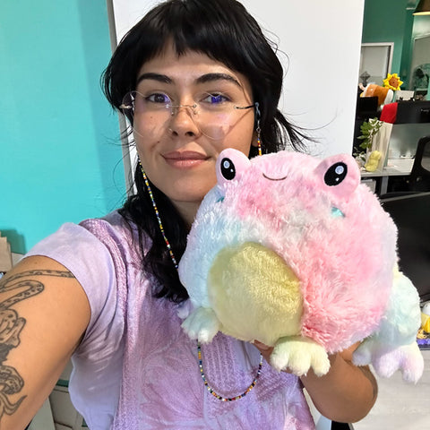 A woman holds the plush tie dye frog near her face. It is slightly larger than her head.