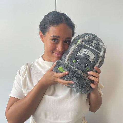 Image shows a woman holding the stuffed tombstone up to her face. It is slightly larger than her head.