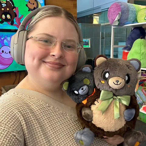 A woman wearing headphones holds the plush two-headed bear. It is slightly larger than her head.