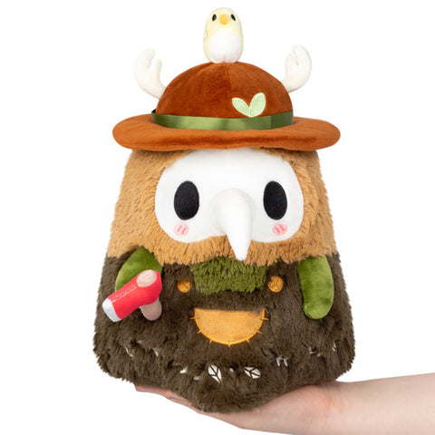A plush toy resembling a woodland version of the Plague Doctor with brown, green, and beige colors, holding a small bird on its head. It is held in someone's hand.