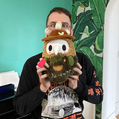 A man holds the plush woodland plague doctor near his face. It is slightly larger than his head.