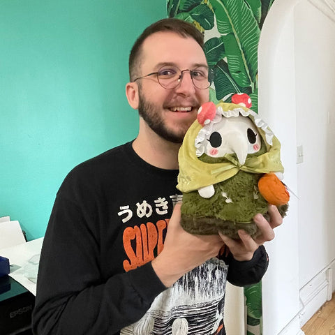 A man holds the plush woodland Plague Nurse near his face. It is slightly larger than his head.