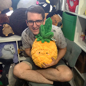 A man holds the plush pineapple in his lap. It is about the size of his torso.