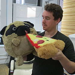 Image shows a man feeding the stuffed pug a piece of stuffed pizza. The pug is much larger than his head.